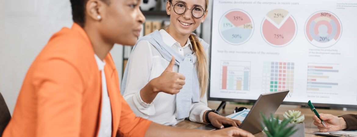 Caucasian woman in eyeglasses smiling on camera showing thumb up while sitting with female african colleague at desk. Two economists spending time at office for work on financial report.