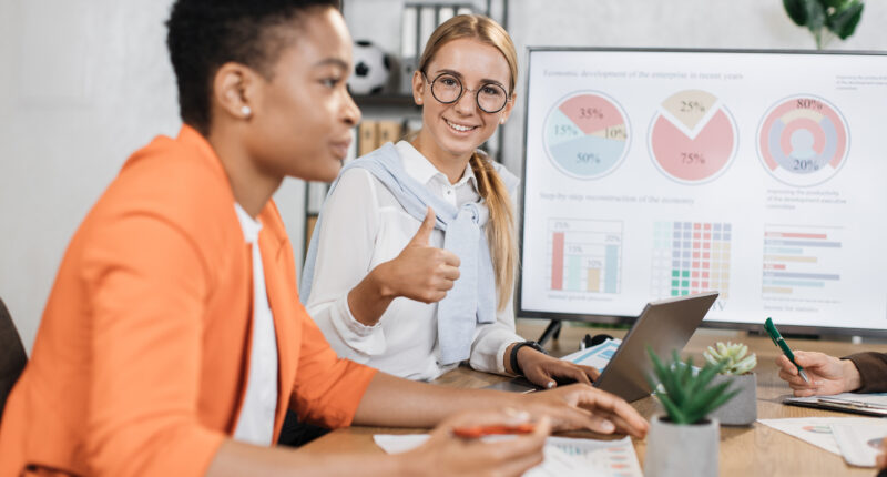 Caucasian woman in eyeglasses smiling on camera showing thumb up while sitting with female african colleague at desk. Two economists spending time at office for work on financial report.